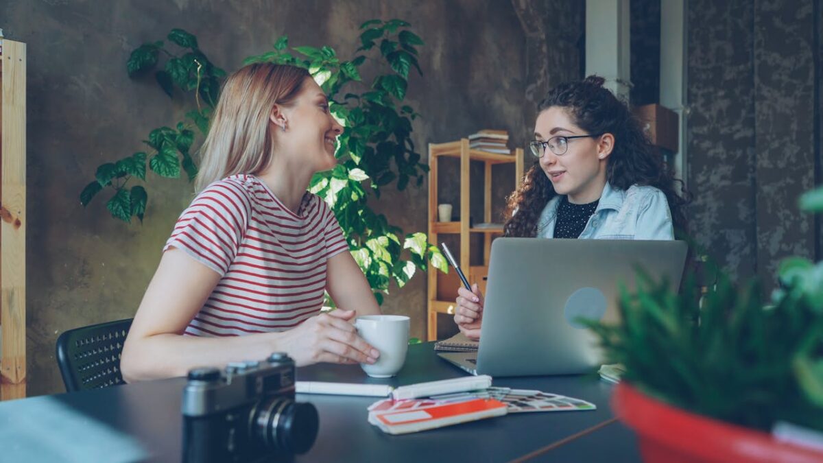 Two women discussing ideas at a laptop workspace with coffee.