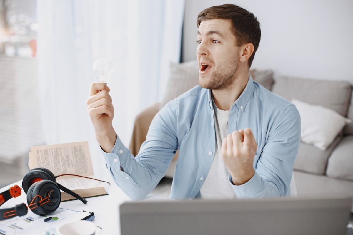 Man joyfully holding a light bulb, symbolizing creativity and innovation in a home office setting.